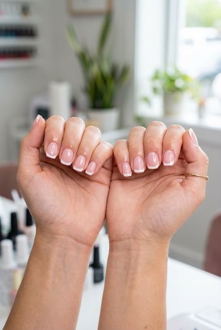 A close-up of two hands showcasing a stylish nude french manicure with delicate nail art, set against a bright and modern salon background.