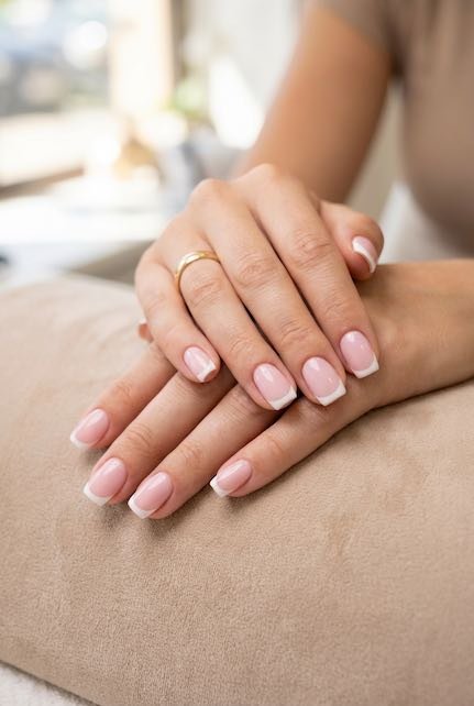 Close-up of neatly manicured hands featuring a pink French manicure and a delicate gold ring, resting on a soft surface.