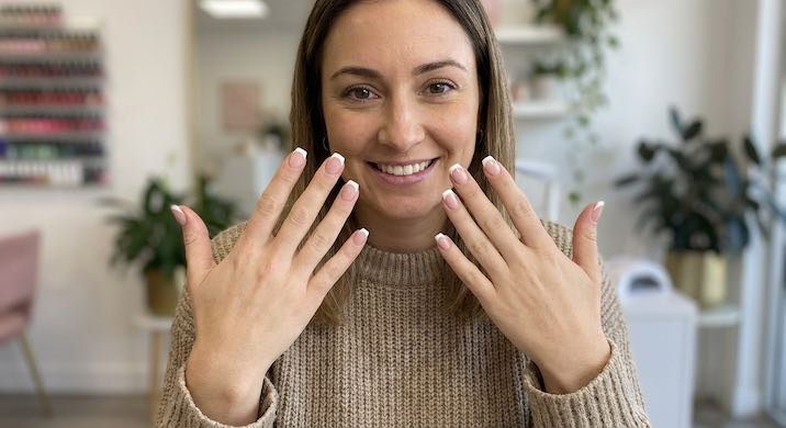 A woman smiling and showing off her manicured hands with elegant French tip nails in a nail salon setting.
