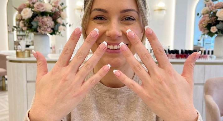 A smiling woman shows off her beautifully manicured hands with pink and white nails in a stylish salon setting.