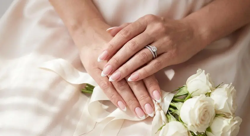 1wed - PolishedAndPrettyNails.com Classic bridal French manicure with white tips and a wedding ring, hands resting on a white rose bouquet